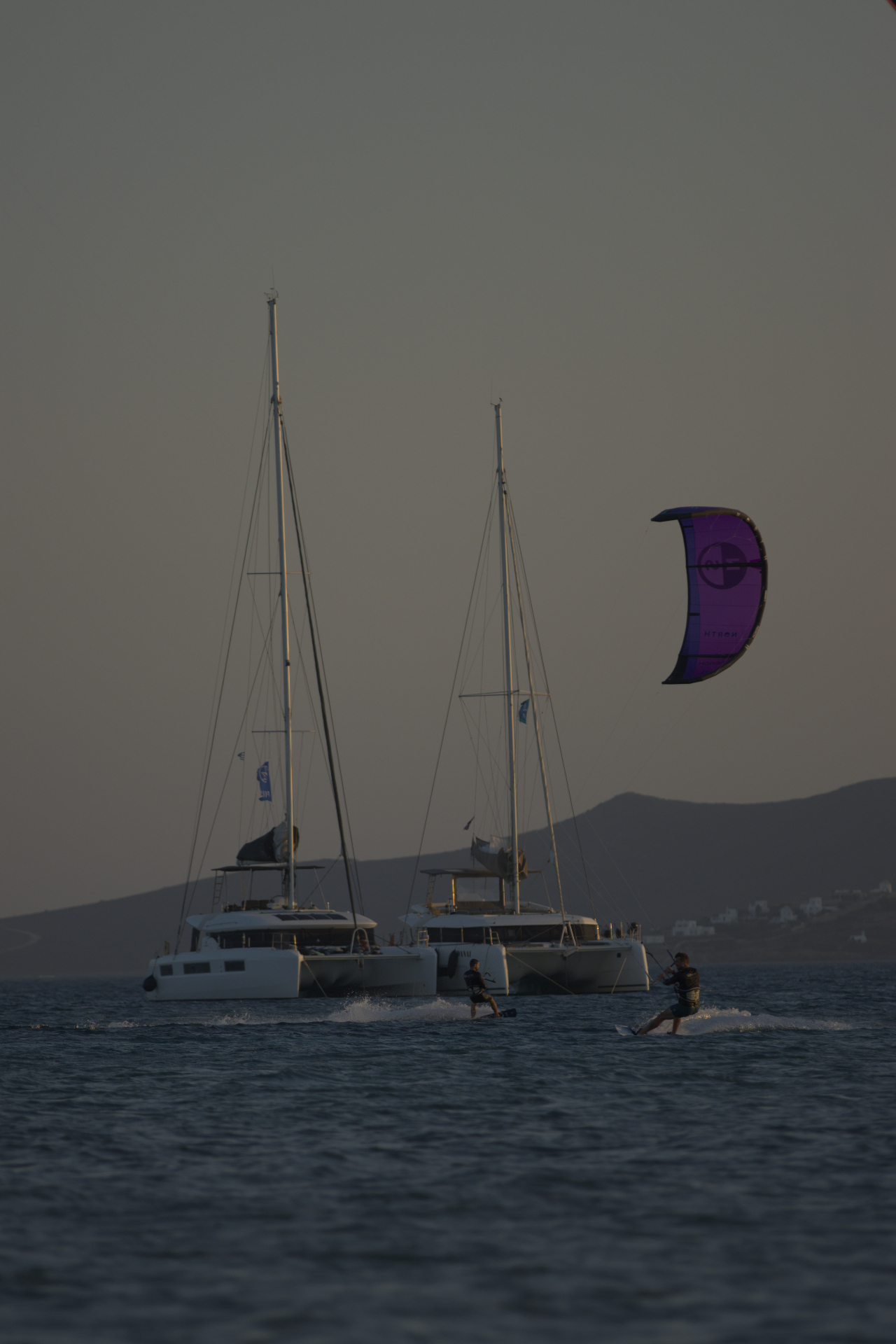 Riders crossing in front of two anchored catamarans at dusk