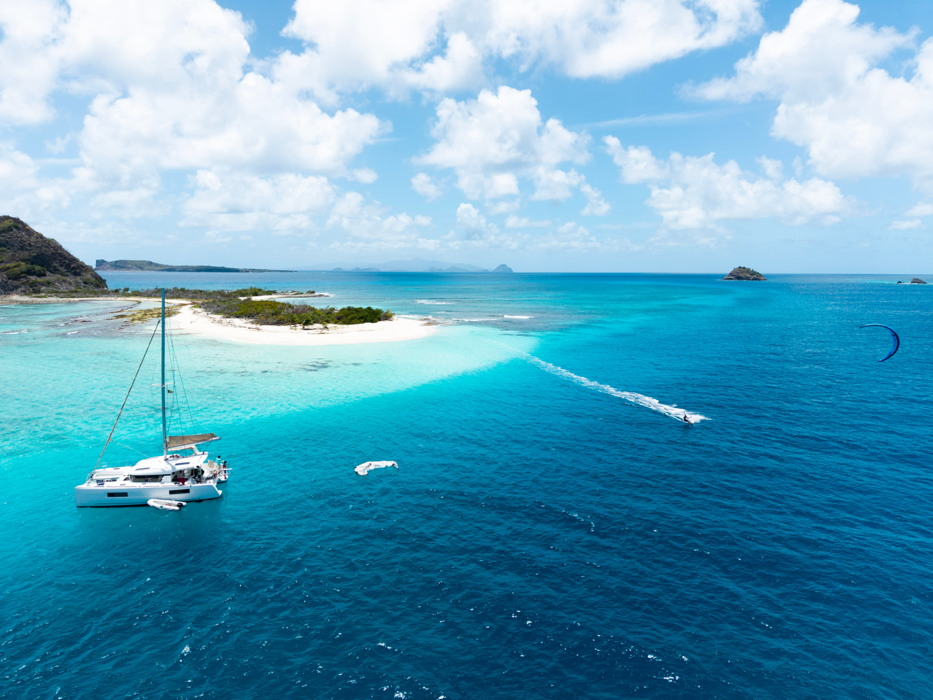 Drone view of the catamaran anchored off a white-sand cay as a kiter carves across deep blue water